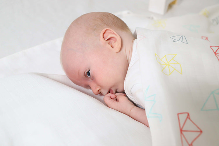 Baby facing down on white sheet, covered in printed musluv as a blanket