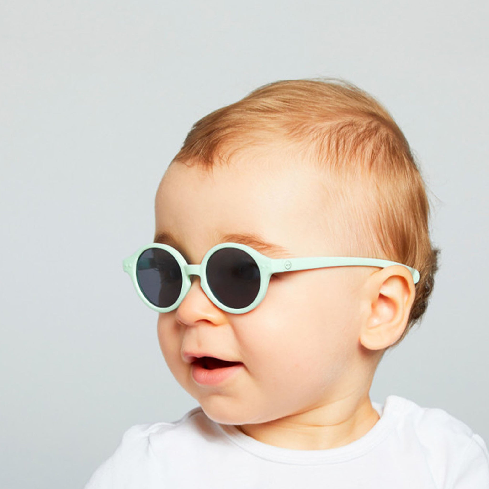 Baby wearing sky blue sunnies looking towards left off screen with grey background 