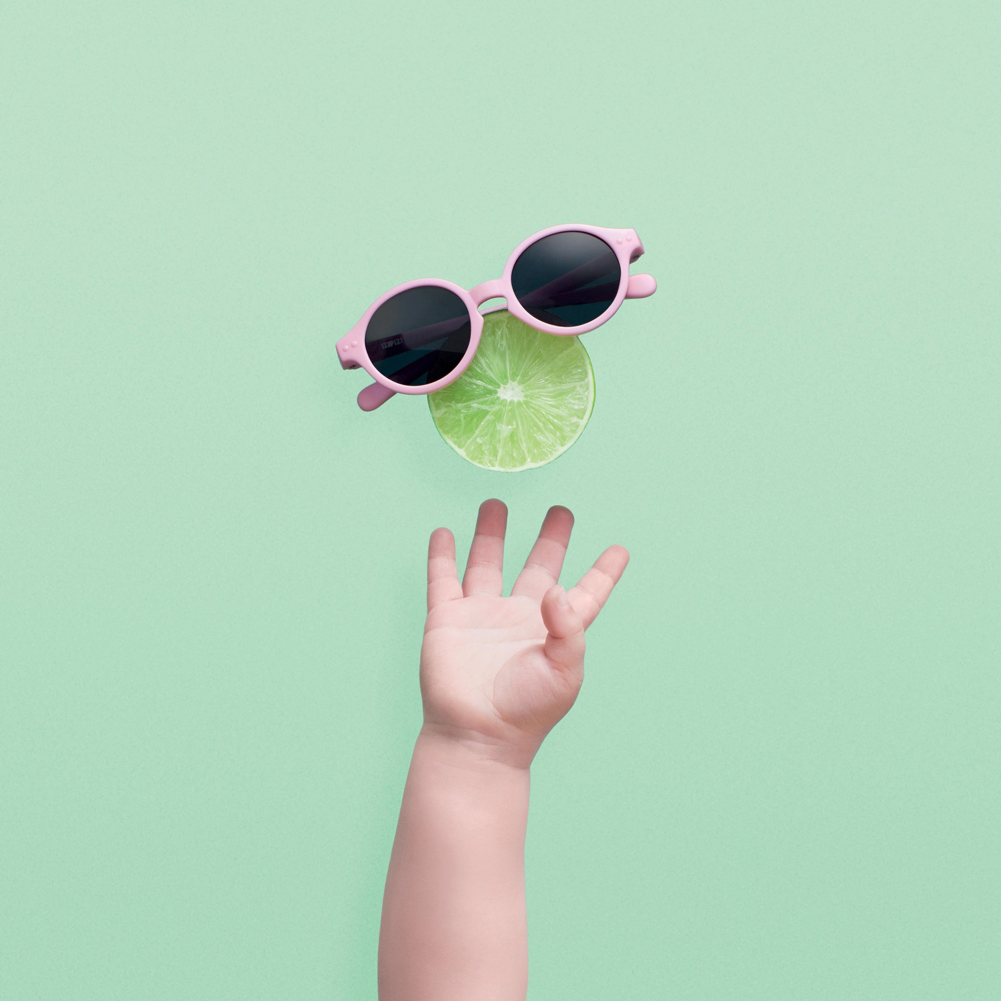 Baby's hand reaching for a half a lime in the air, with pink sunglasses perched on top of lie. Green background 